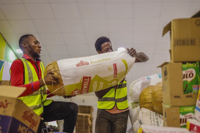 Two male volunteers organizing large sacks and cardboard boxes of relief goods indoors.